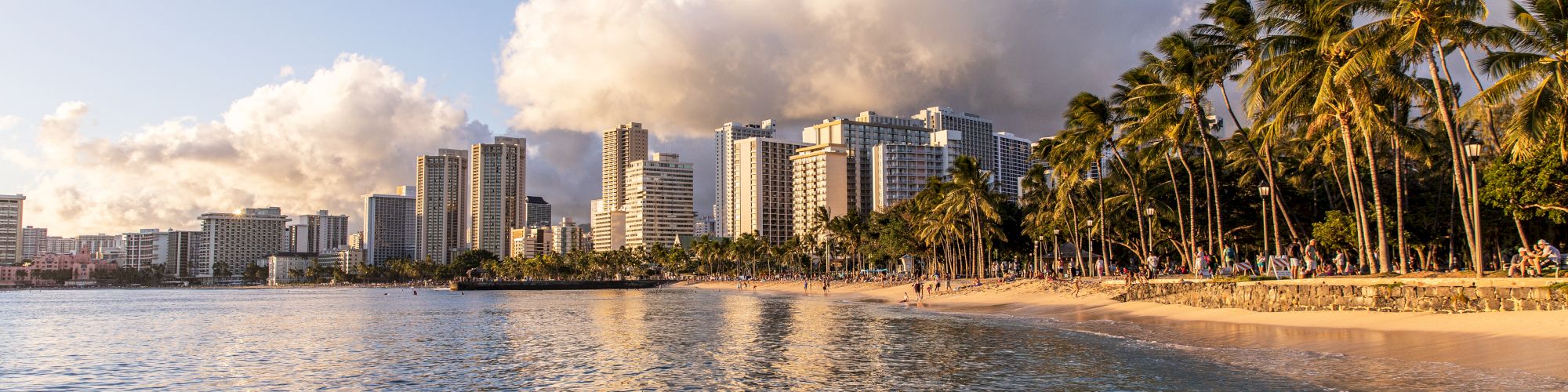 A tropical coastal city with a sandy beach, palm trees, and calm blue water, high-rise buildings skyline in the background under a partly cloudy sky.