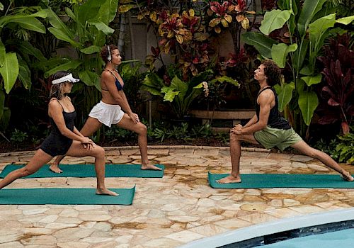 Three people are practicing yoga on mats near a pool, surrounded by lush greenery and tropical plants.