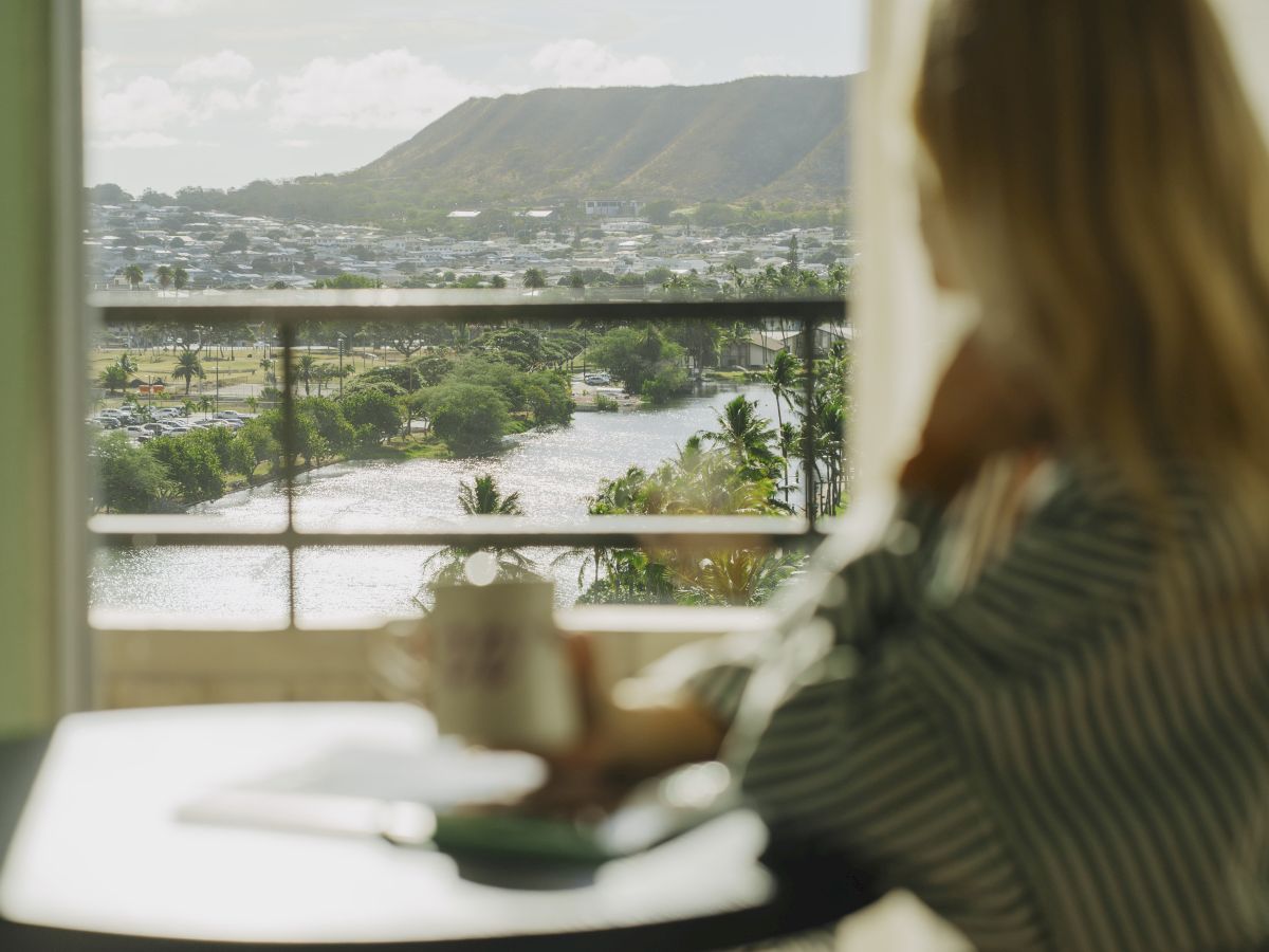 A person sits at a table indoors, looking out a window at a river and hills in the distance, blurred in the foreground.