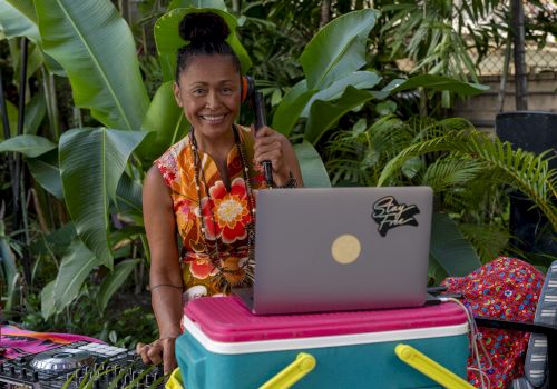 Smiling woman DJing outdoors with a laptop and microphone, surrounded by big green tropical leaves and colorful cushions.