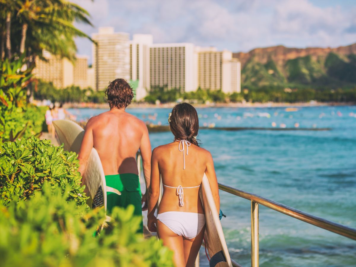 Two people in swimwear walk along a beachfront promenade toward the water, with palm trees, hotels, and mountains in the background.