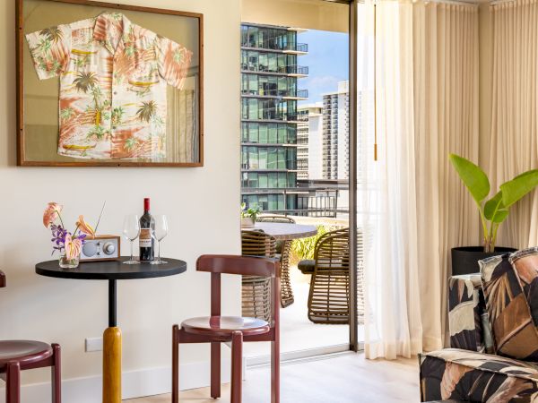 A cozy room with a small table and two chairs, tropical decor, a framed shirt, wine, and a parrot statue, leading to a balcony with city views.