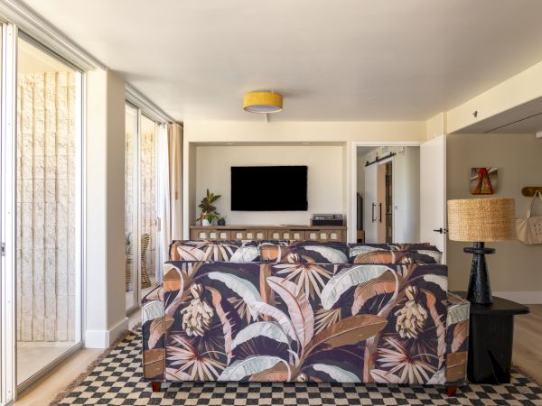 A living room with a patterned sofa, wall-mounted TV, sliding glass doors, and a checkered rug. The room has a cozy and modern decor style.
