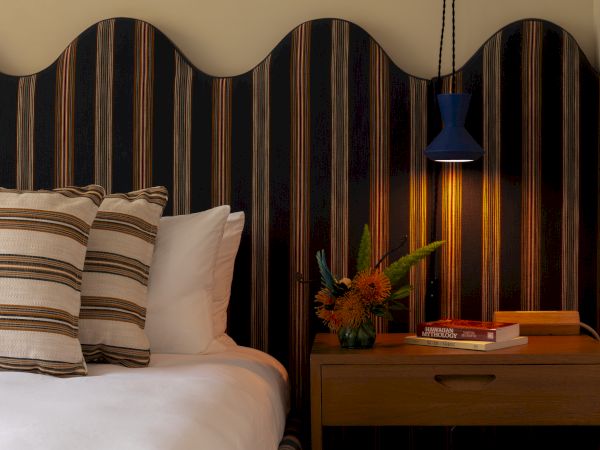 A cozy bedroom setup with a bed featuring striped pillows, a wooden nightstand with a flower vase, books, and a lamp in a blue shade hanging above.