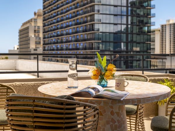 A rooftop patio with a round table featuring a magazine, a vase of flowers, a carafe of water, and a mug. Buildings are in the background.