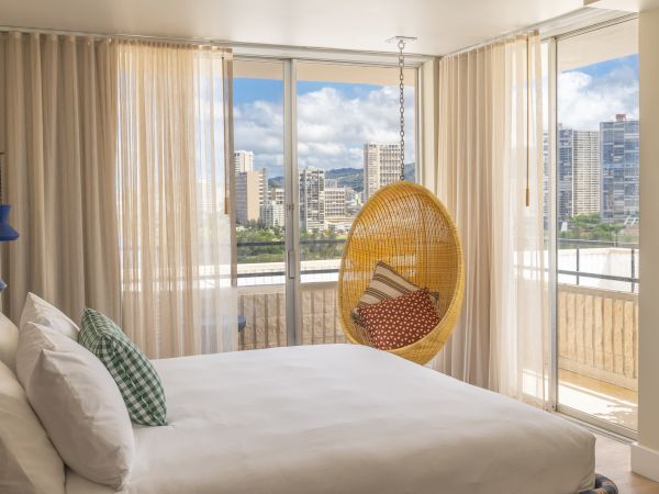A modern bedroom with a large bed, hanging chair, and floor-to-ceiling windows showing a cityscape view, with sheer curtains partially drawn.