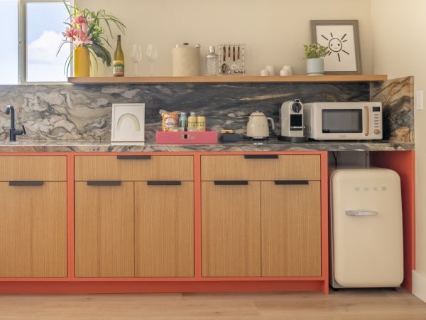 The image shows a modern kitchen with wooden cabinets, a marble backsplash, and various small appliances and decor, including a microwave and a mini-fridge.