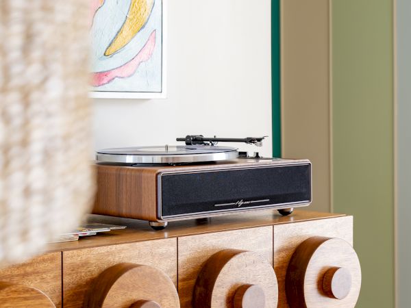 A modern vinyl record player sits atop a stylish wooden cabinet with circular patterns; a colorful abstract painting hangs above it.