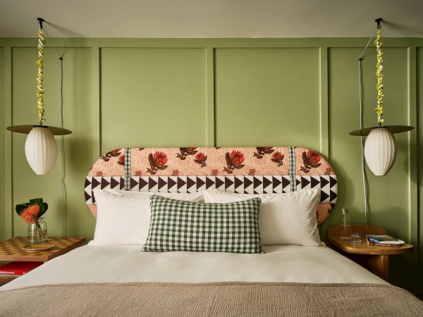 A neatly made bed with patterned headboard, flanked by hanging lamps and nightstands, decorated with a green plaid pillow and flowers.
