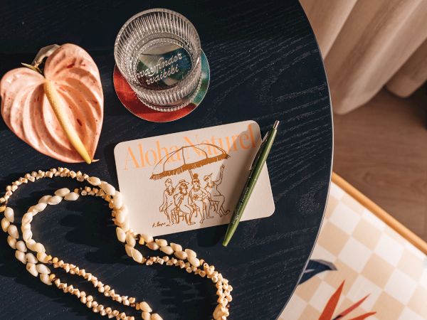 A table with a shell necklace, postcard, pen, tropical plant, and glass on a coaster labeled "Wayfinder Waikiki," in warm lighting.