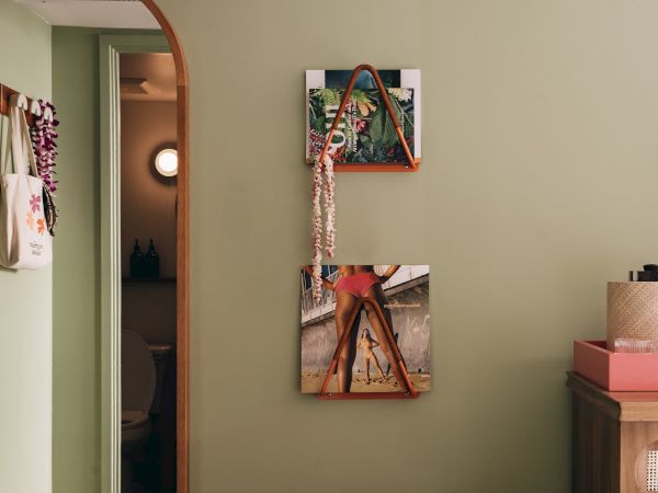 A room with a floral ottoman topped with a straw hat, wall art, and a mirror beside a door to a bathroom with a visible toilet.