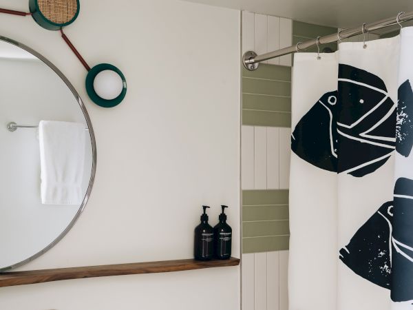 A modern bathroom with a round mirror, wall shelf, two black bottles, decorative shower curtain, and light wood vanity.