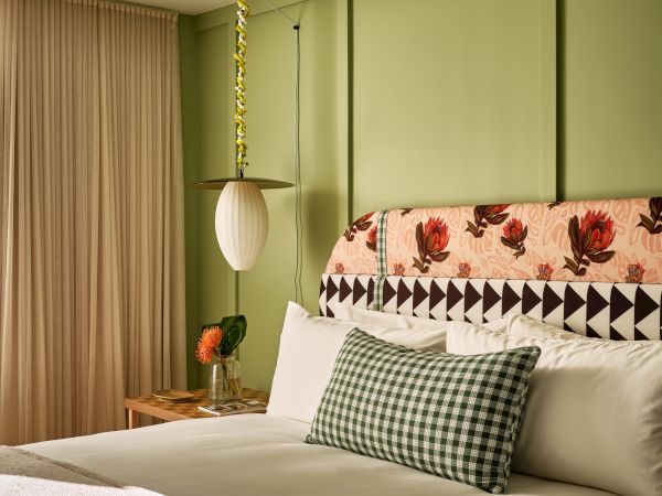 A cozy bedroom with a floral headboard, green wall, patterned pillow, hanging lamp, and side table with flowers and books.