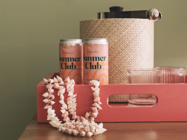 A wooden table holds a coral tray with two "Summer Club" cans, a shell necklace, an ice bucket, and glassware against a green wall.