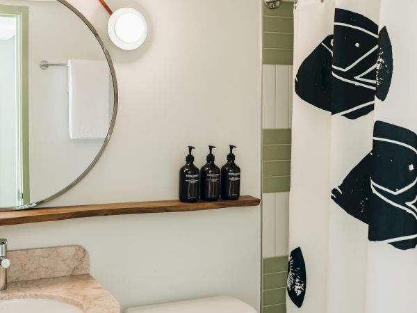 A bathroom with a round mirror, three soap dispensers on a shelf, and a shower curtain featuring abstract fish designs.