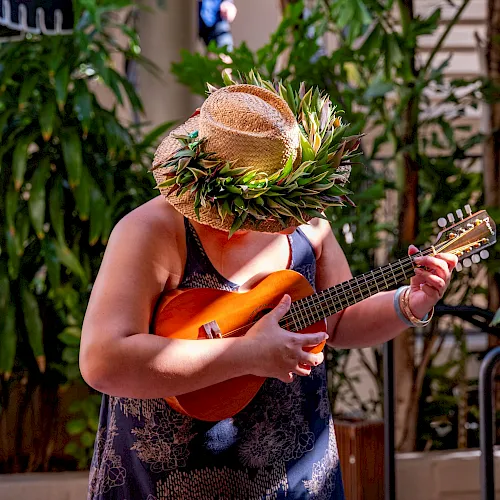 A woman wearing a straw flower hat plays an orange ukulele outdoors in lush green garden.