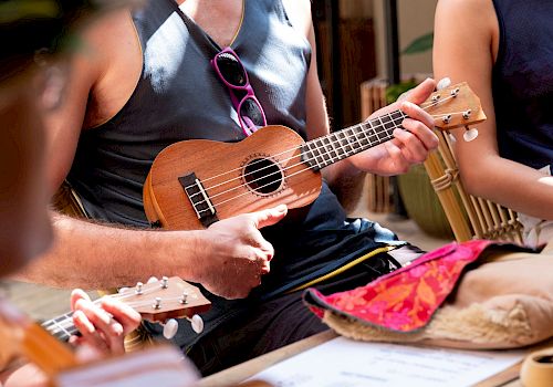 A bearded man in a blue tank top strums a ukulele during a casual music session with others indoors.