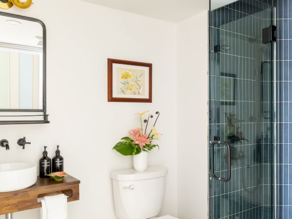 A bathroom with a mirror, sink, and toilet with flowers on top, and a framed picture on the wall next to a glass shower door ends the sentence.