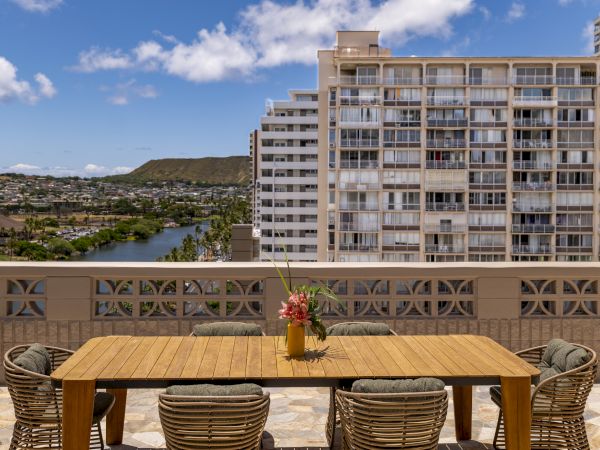 A rooftop terrace with a wooden table and six chairs overlooking a cityscape with buildings, a water body, and hills under a partly cloudy sky.