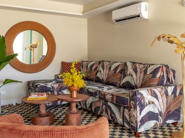 A cozy living room with a patterned sofa, round mirror, potted plants, coffee table, and cushions, adorned with warm tones and tropical decor.