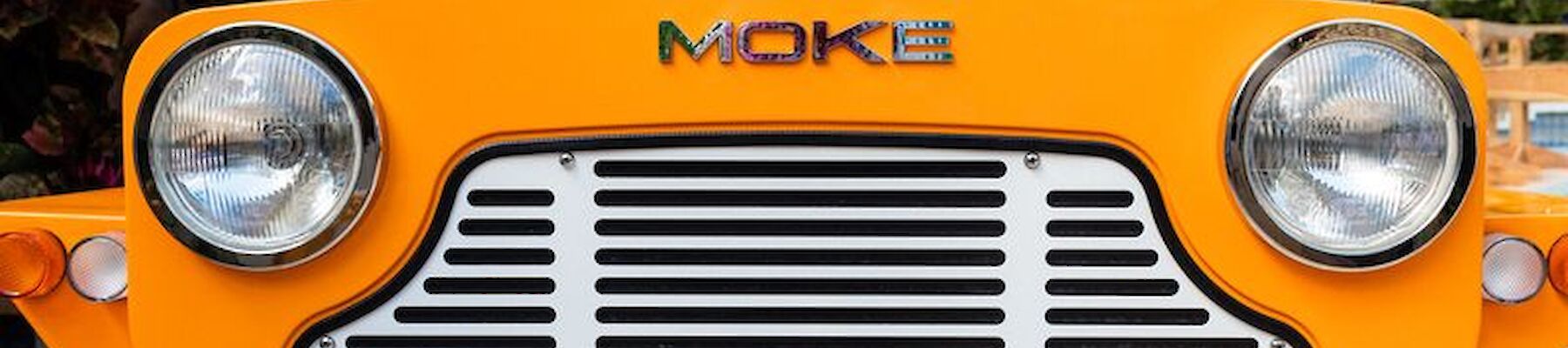A bright yellow vintage-style car with "MOKE" on the front, featuring large headlights and a distinctive grille design.