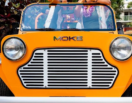 A bright yellow vintage-style car with "MOKE" on the front, featuring large headlights and a distinctive grille design.
