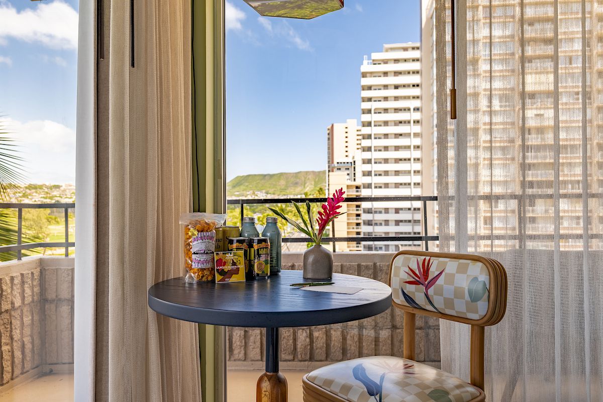 A small balcony with a round table and chair, snacks, drinks, and a vase with a flower, overlooking city buildings on a clear day.