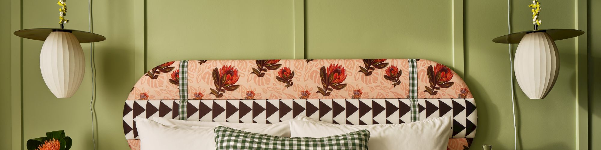 A cozy bedroom with a green paneled wall, patterned headboard, checked pillow, and hanging lamps, featuring side tables with decorative items.