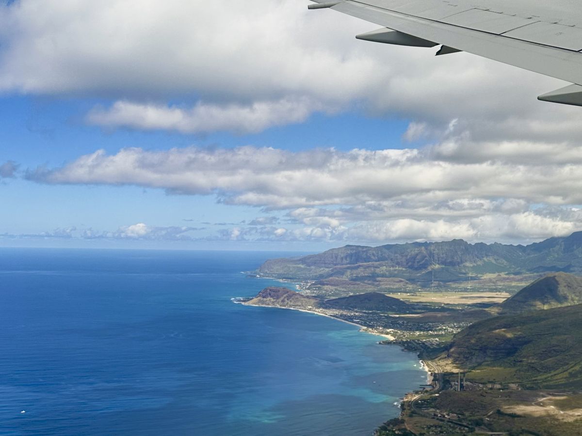 Aerial view of a coastline with turquoise water, green hills, and a few buildings near the shore, seen from an airplane window.