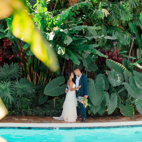 A couple in formal attire embraces by a pool, surrounded by lush greenery in a tropical setting.