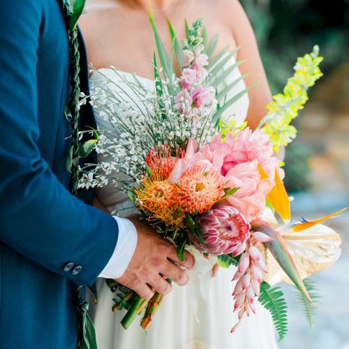 A couple dressed for a wedding, the bride holding a colorful bouquet with pink and yellow flowers, standing close together outdoors.