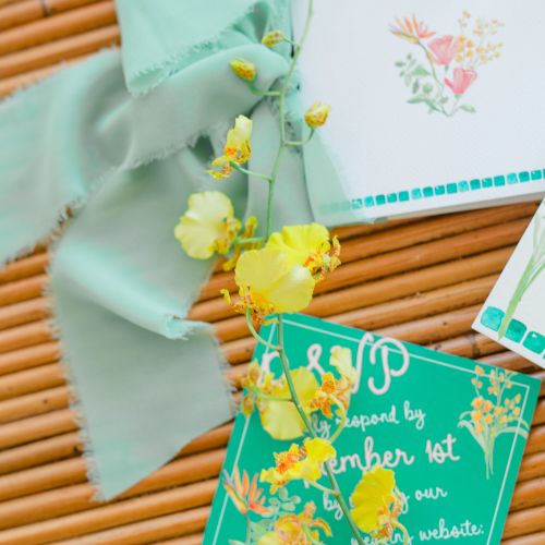 The image shows a vow card with flowers, a ribbon, and a teal RSVP card with yellow flowers, on a wooden surface.