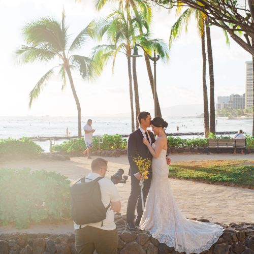 A couple in wedding attire poses by the beach with palm trees, while a photographer captures the moment.