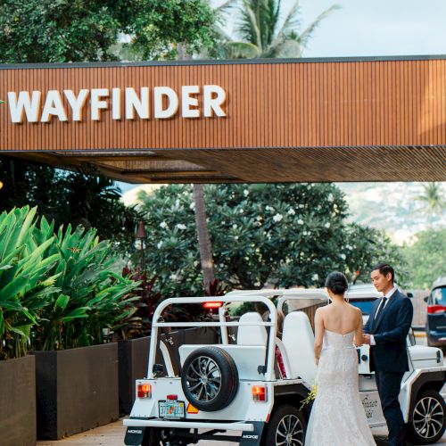 A couple in formal attire stands beside a white open-top vehicle under a sign reading "Wayfinder," surrounded by lush greenery.