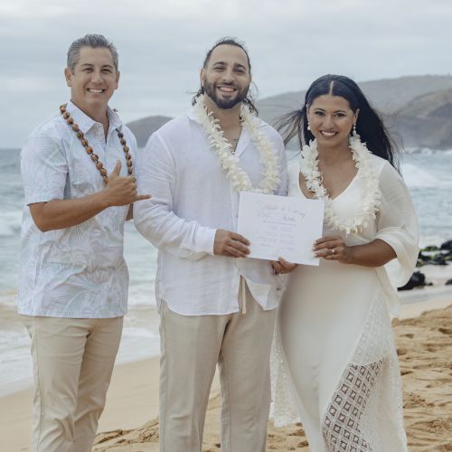 Three people on a beach; two are celebrating with a certificate. They're wearing leis and beach attire, with ocean and mountains in the background.
