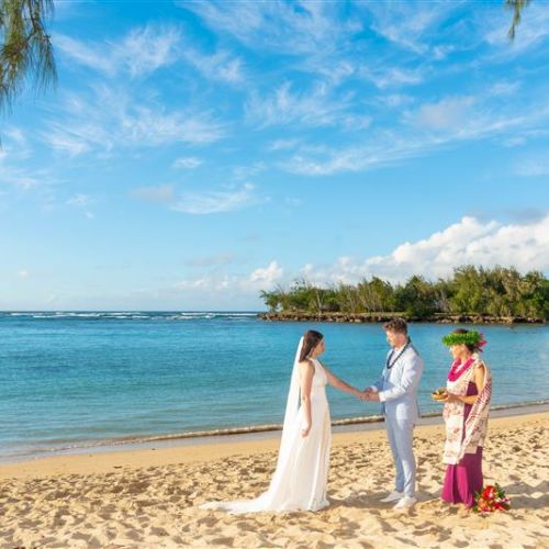 A couple is getting married on a sandy beach, with a person officiating the ceremony against a backdrop of ocean and clear skies.