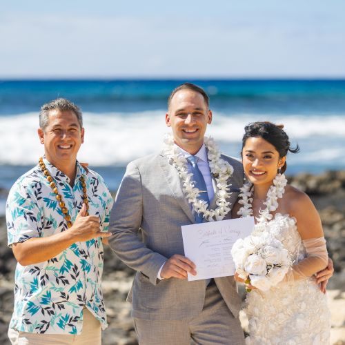 A couple in wedding attire stands on a beach with a man in a Hawaiian shirt; they're holding a certificate, with the sea in the background.