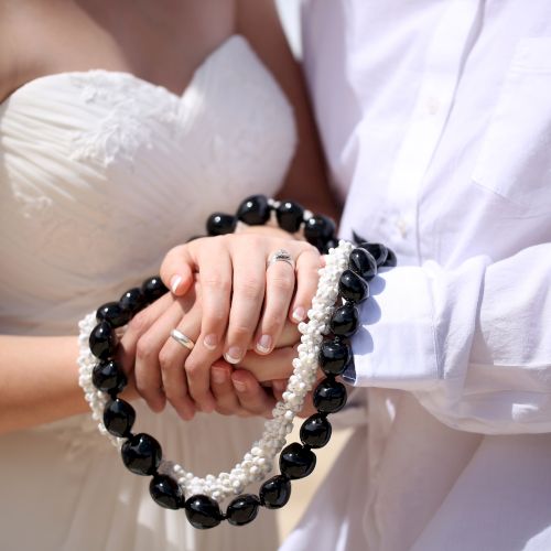 A couple in wedding attire holds hands with a lei or garland of black and white beads draped over them.