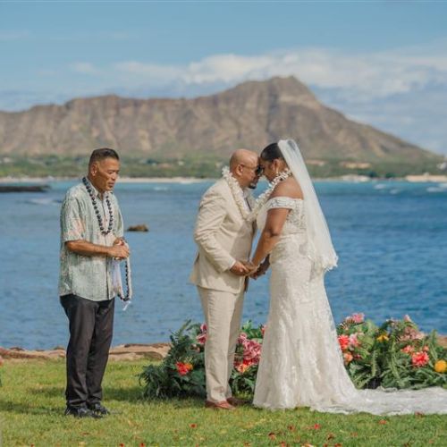 A couple getting married outdoors by the ocean, with a mountain and cityscape in the background, surrounded by colorful flowers and guests.