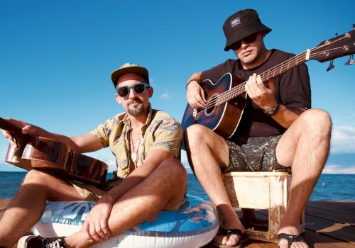 Two men sit outdoors by the water, playing guitars. They wear hats and sunglasses, embracing a casual, relaxed vibe under a clear sky.