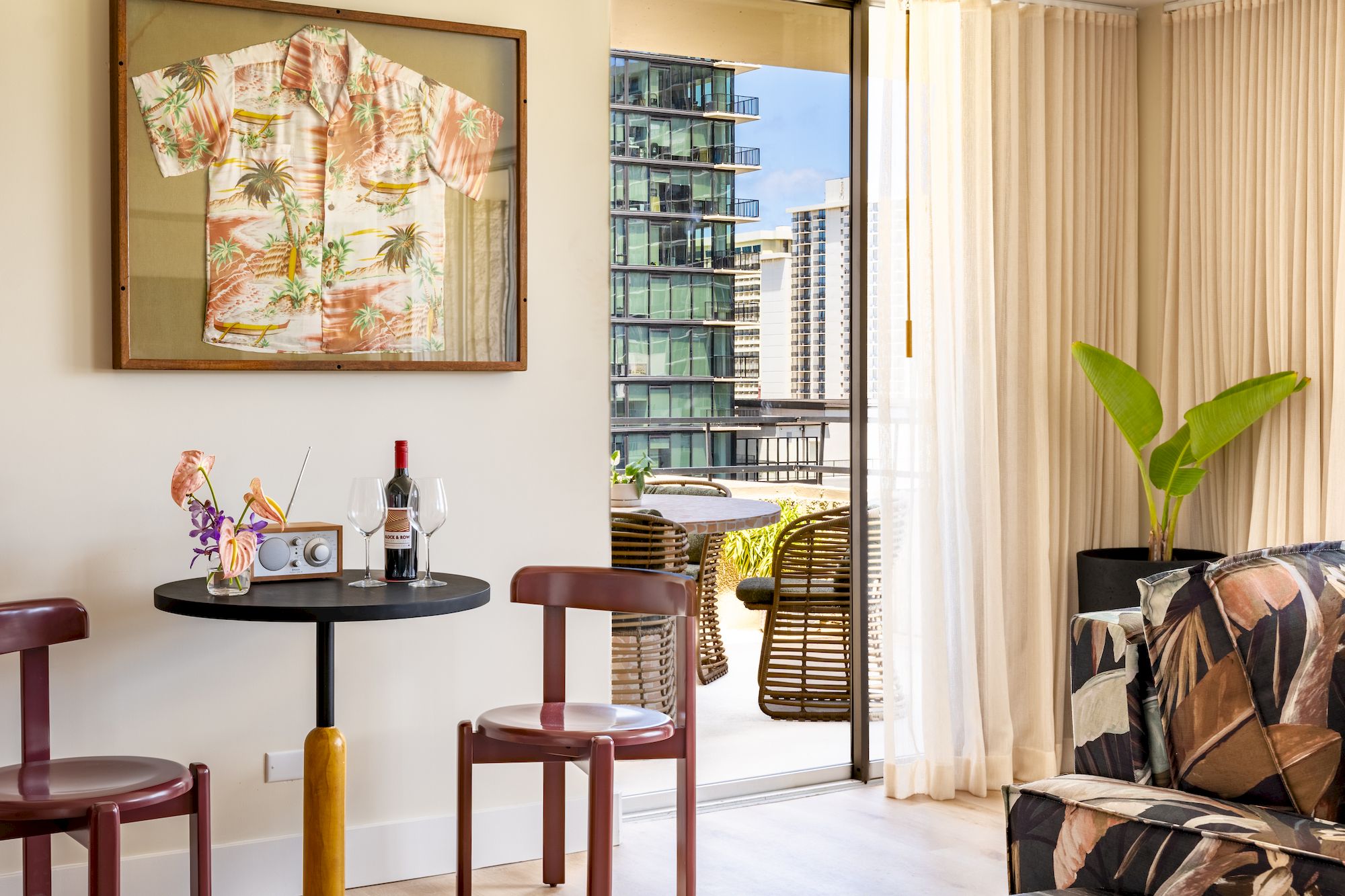 A stylish apartment with a small dining table, two chairs, tropical shirt framed on the wall, a bottle of wine, flowers, and a sunny balcony view.