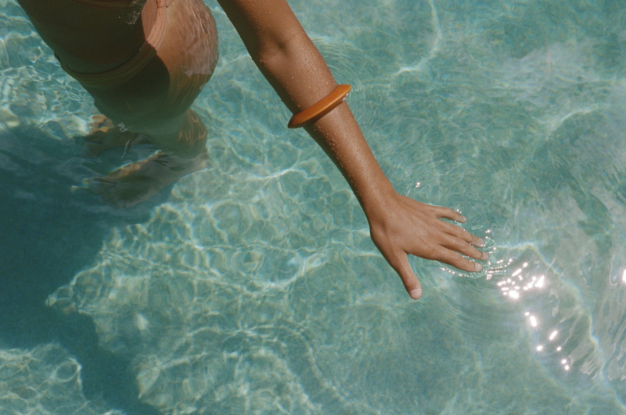 A person paddling or touching clear blue water with their arm and hand, wearing a brown bracelet; sunlit ripples surround the arm.