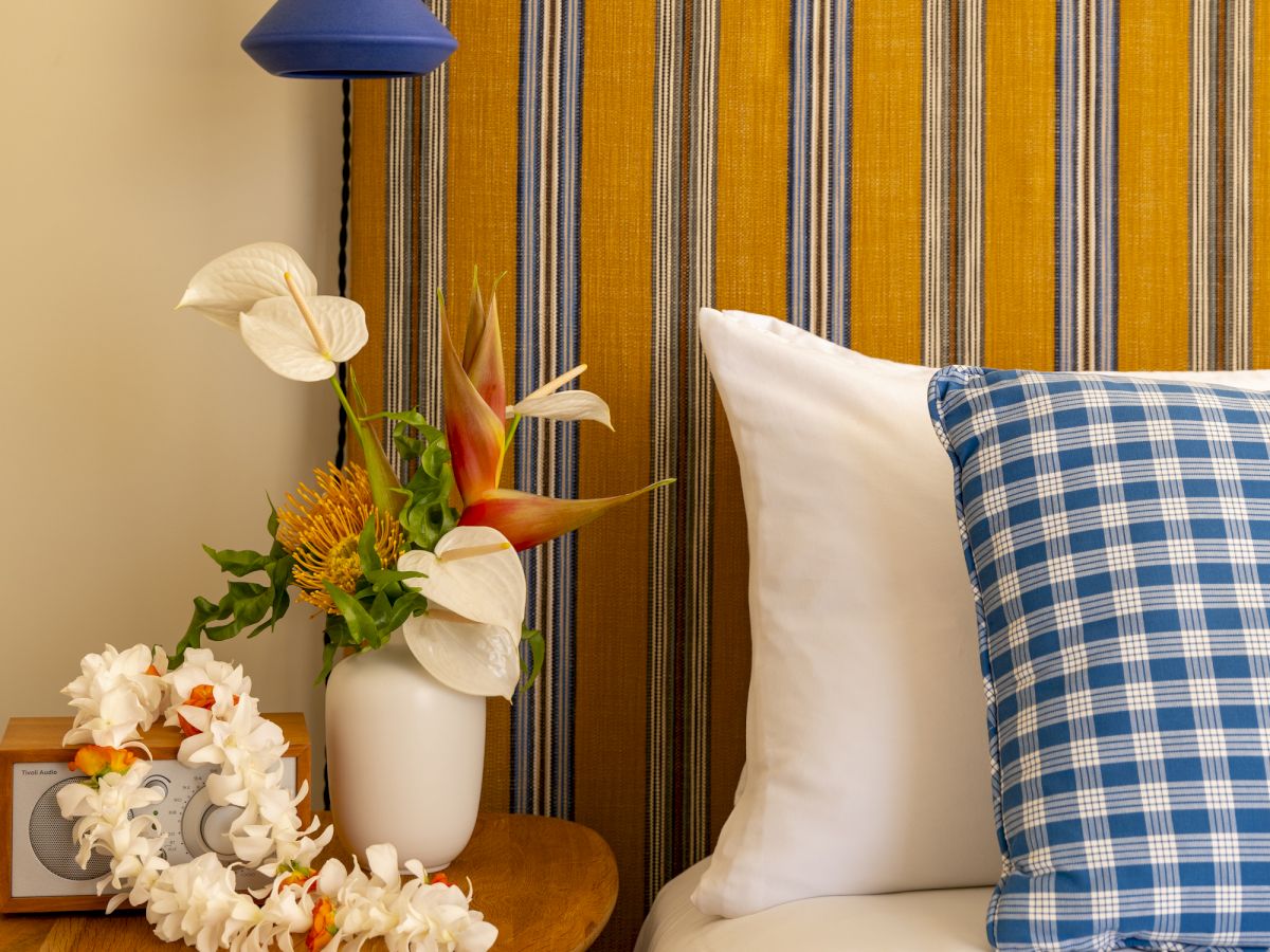 A cozy bedside nook with a striped headboard, blue pendant light, a white vase of flowers, and a blue-checked pillow on a white bed, charmingly arranged.
