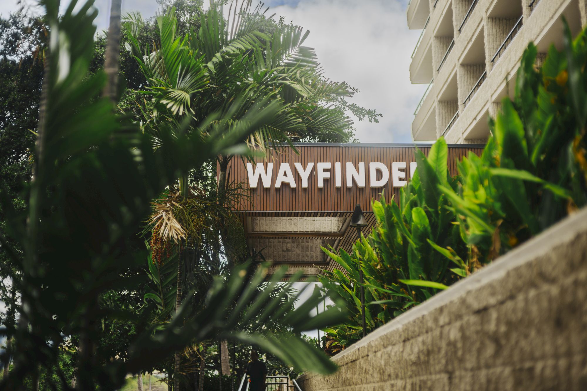 Sign reads &ldquo;WAYFINDER&rdquo; amid lush tropical plants and a concrete wall beside a multi-story building, suggesting a resort or hotel entrance.
