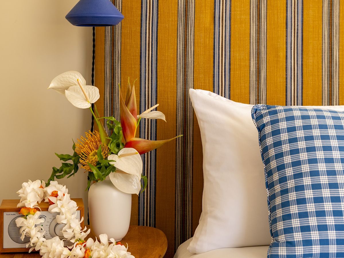 A cozy bedside scene with a striped upholstered headboard, a blue wall sconce, white flowers, and a blue-checked pillow on a white bed.