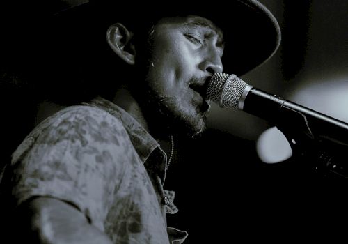 A person is singing into a microphone, wearing a hat, in a dimly lit setting. The image is in black and white.