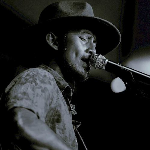 A person is singing into a microphone, wearing a hat, in a dimly lit setting. The image is in black and white.