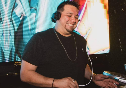 A DJ is smiling while wearing headphones, standing in front of colorful lights, and using equipment.