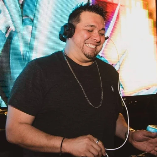 A DJ is smiling while wearing headphones, standing in front of colorful lights, and using equipment.