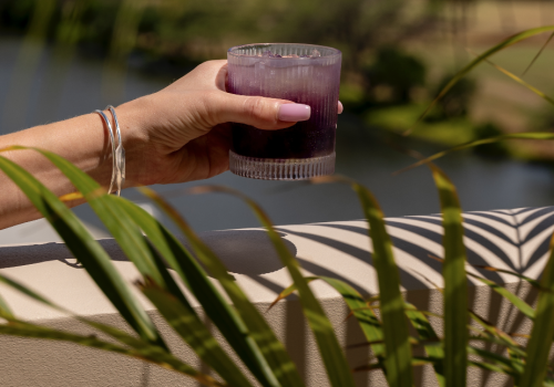 A hand holds a purple drink in a glass against a backdrop of greenery and buildings, viewed through palm leaves on a sunny day.